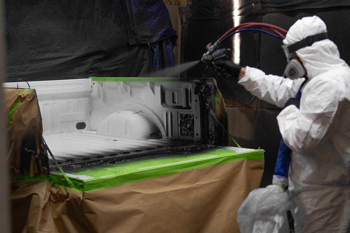 Technician in protective suit sprays black bed liner onto white pickup truck bed.