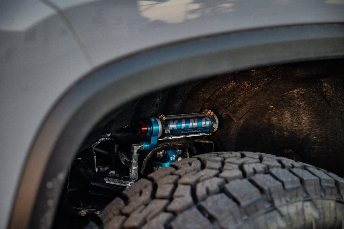 Close-up of a light-colored SUV's off-road suspension featuring a blue King Shocks reservoir and a knobby tire.